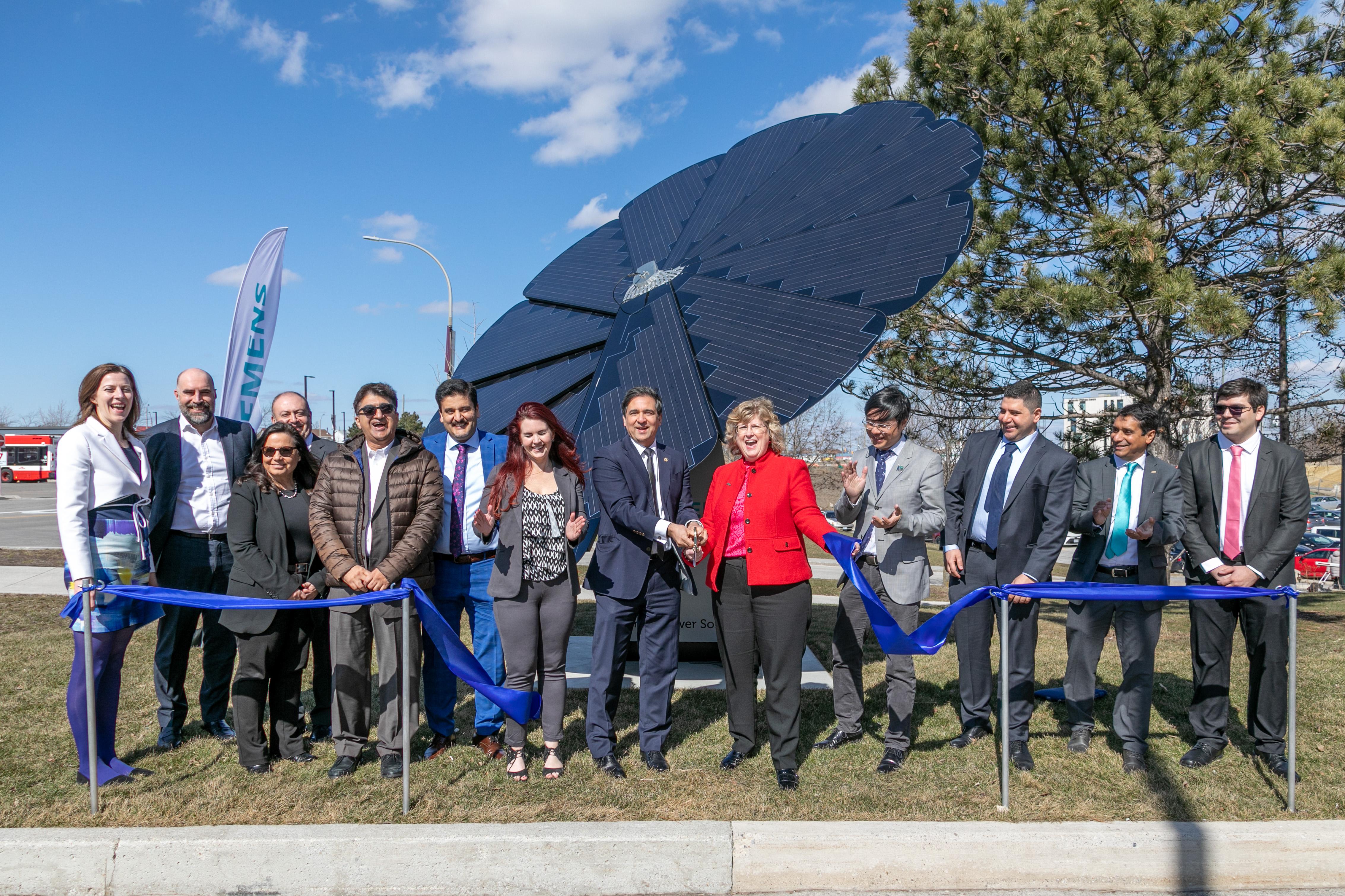A group of people smile and clap as a ribbon is cut. The group is standing in front of a device equipped with solar panels shape