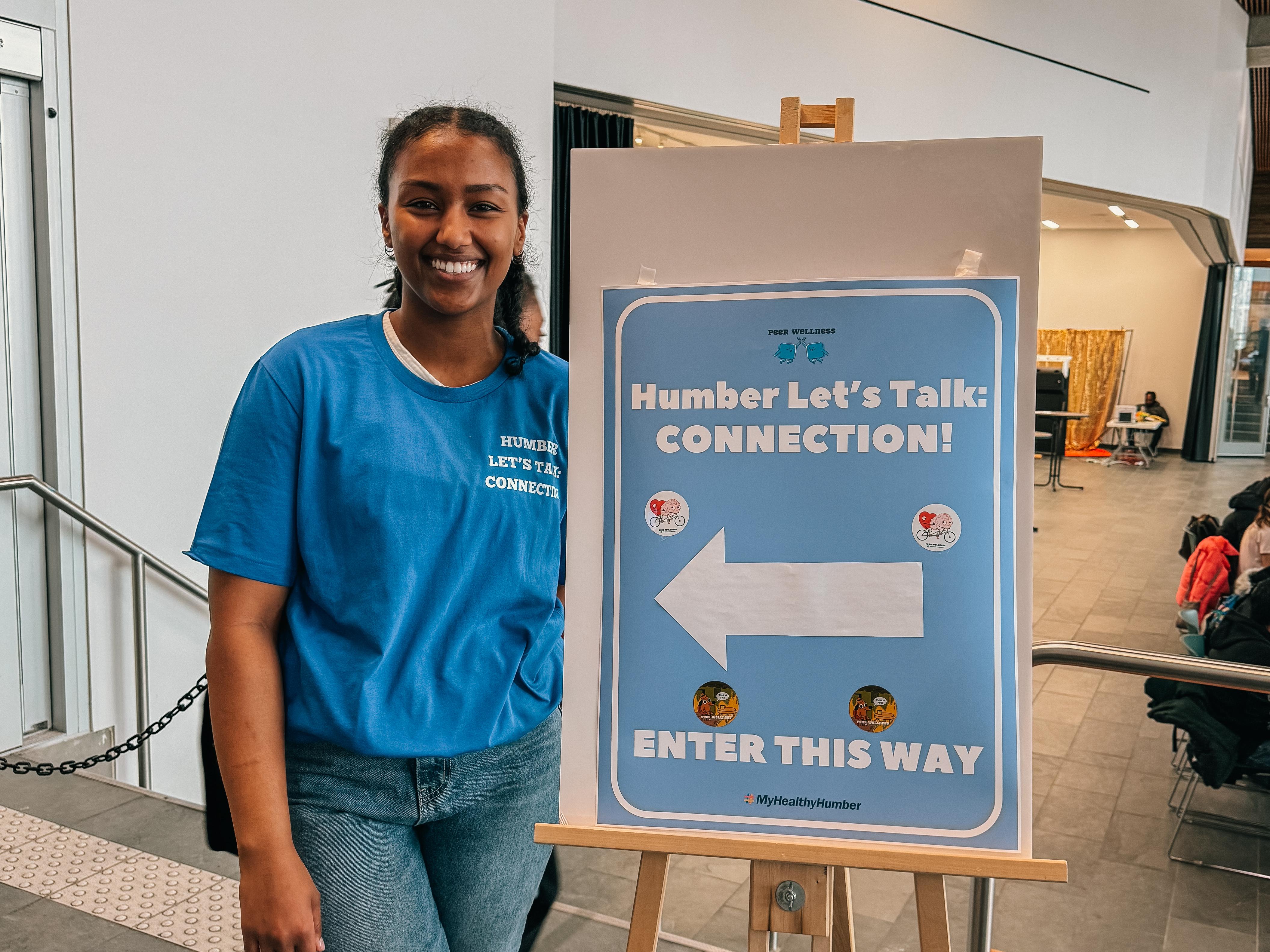 A smiling person stands next to a sign that reads Humber Let’s Talk.