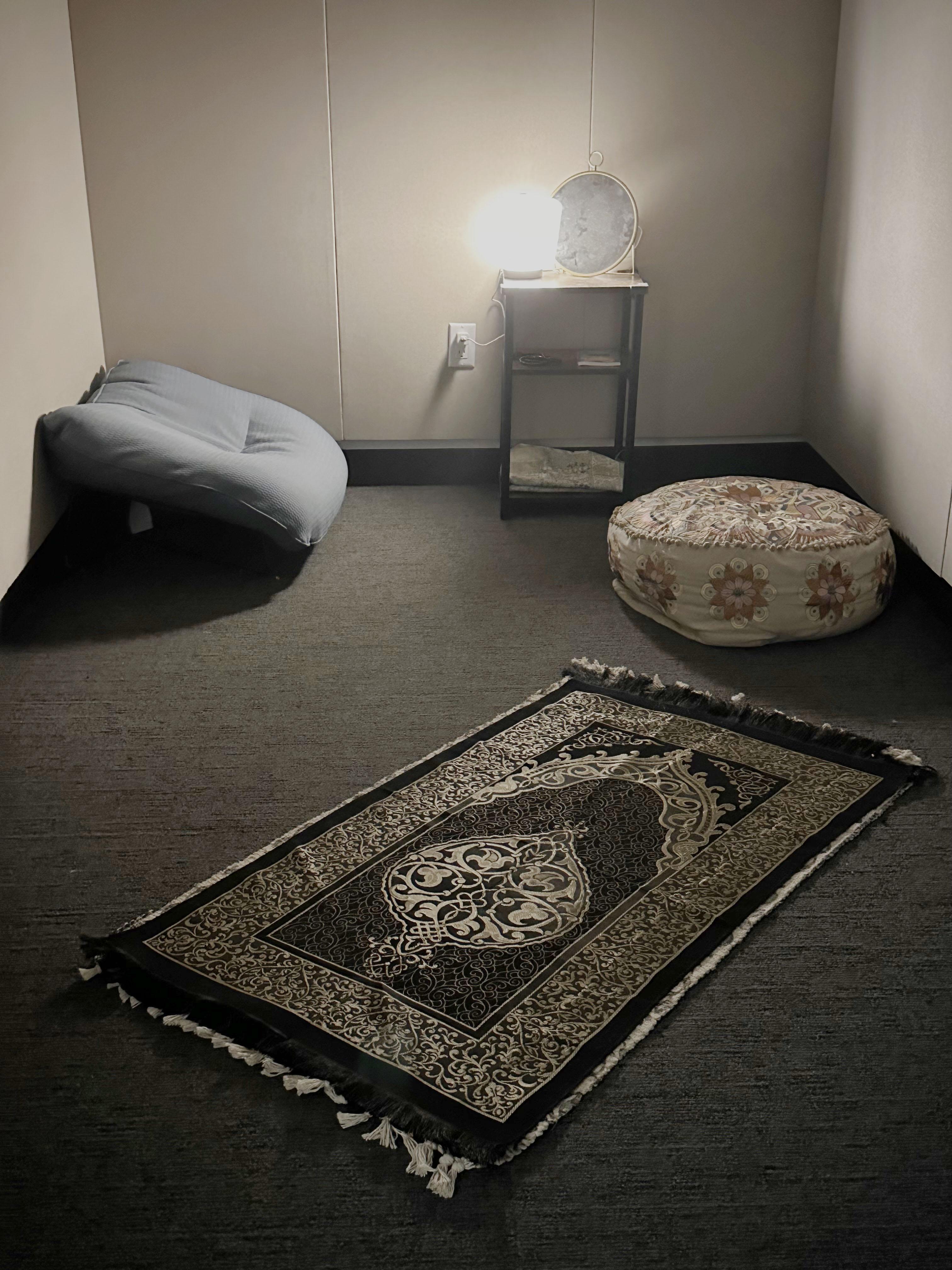 Cushions along with a prayer rug in a room that also has a light sitting on a small table.