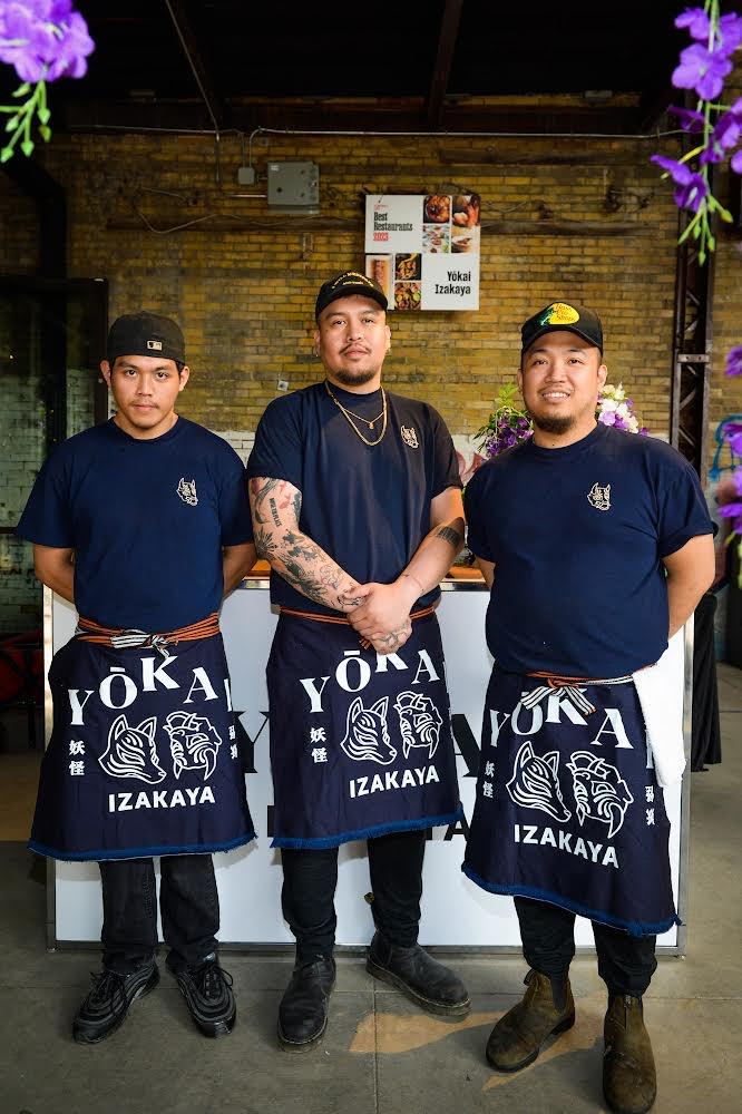Three people stand in a row wearing aprons that read Yokai Izakaya.