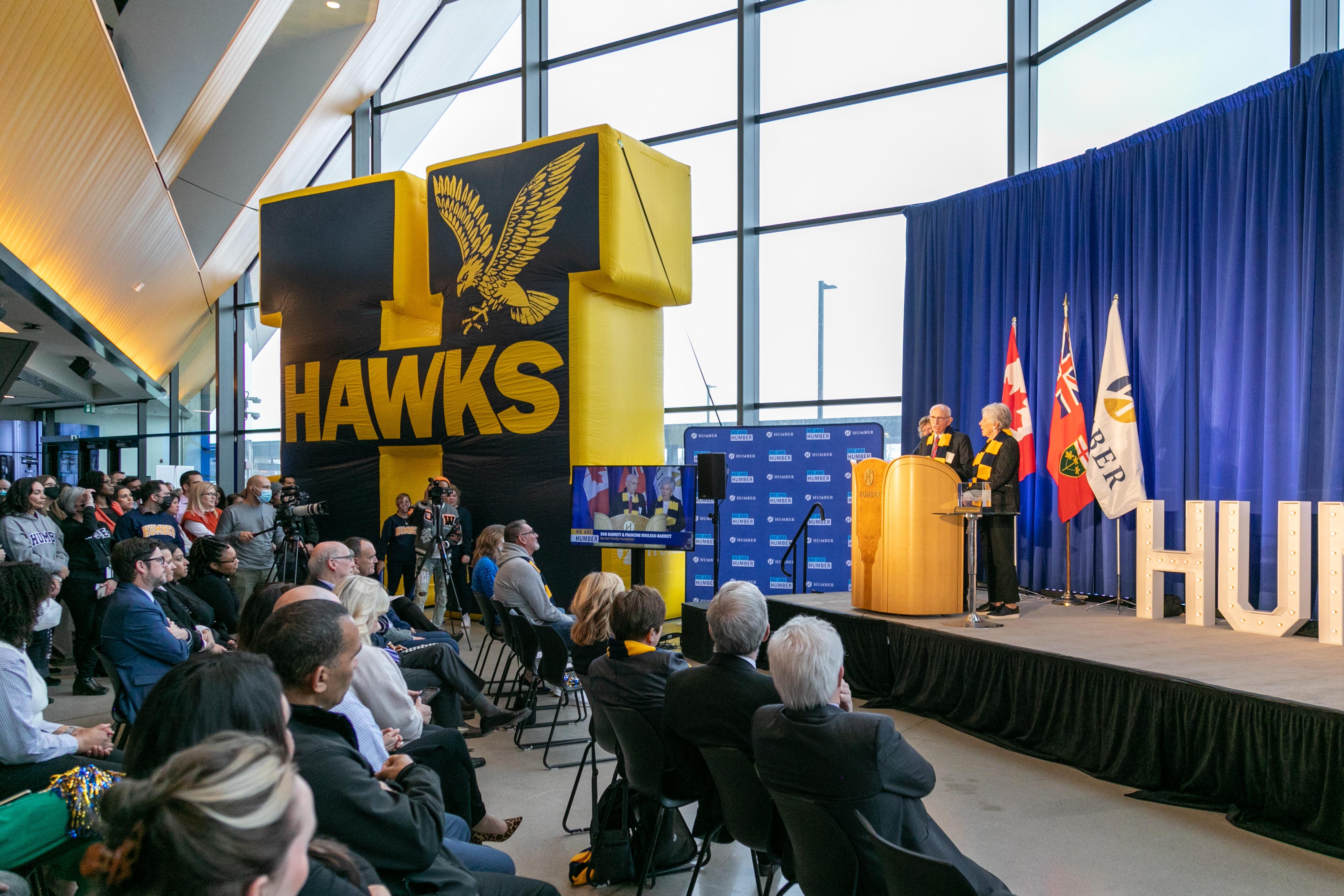 Several people stand at the podium on a stage while a crowd watches and listens.