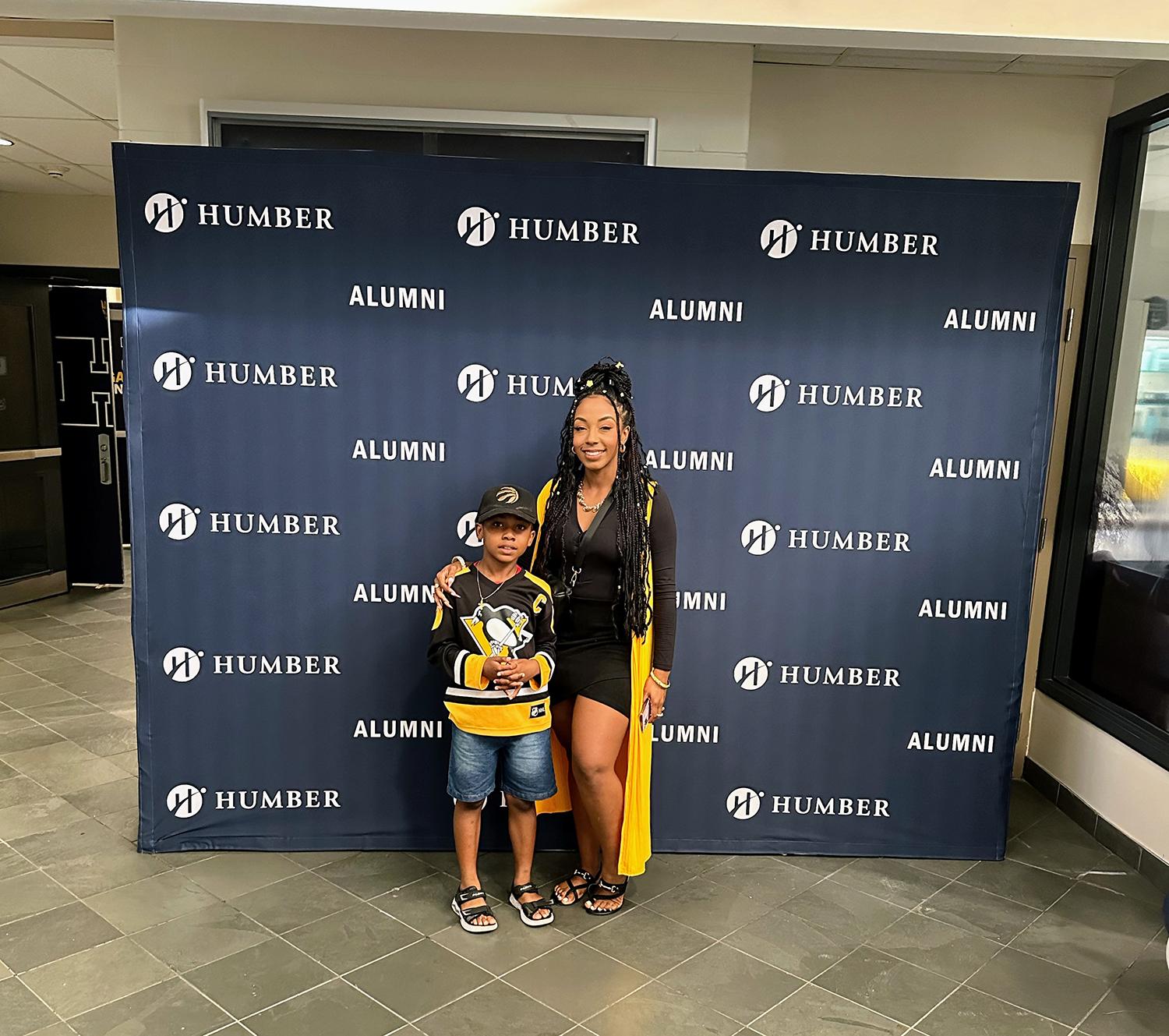 Two people stand together in front of a banner that reads Humber Alumni.