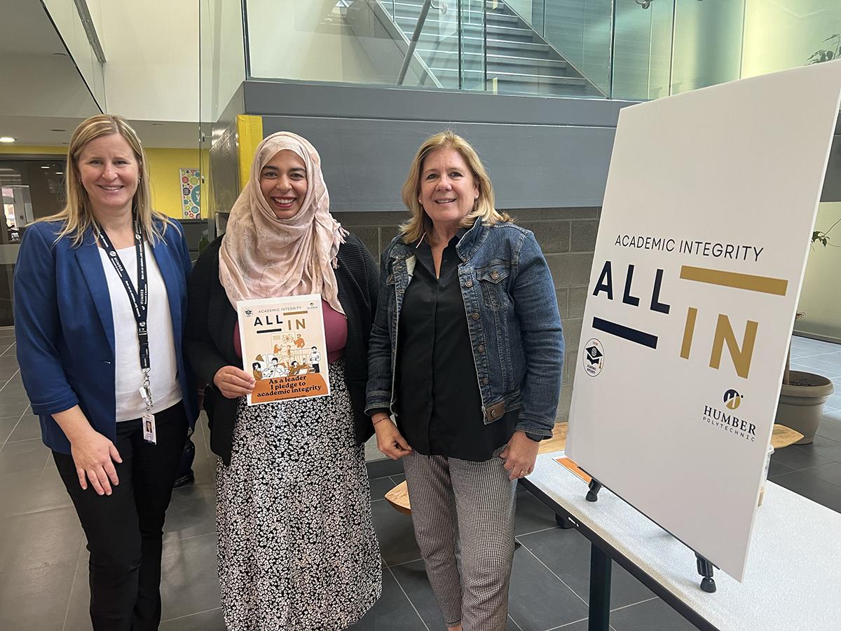 Three people stand together next to a sign that reads Academic Integrity All In.