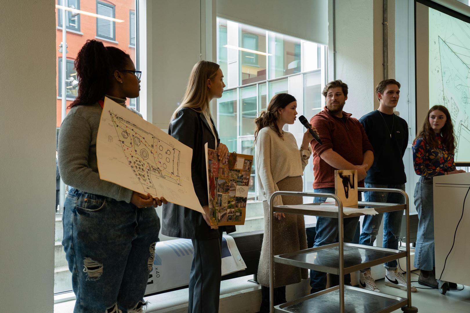 Six students give a presentation. Two are holding up drawings while a third speaks into a microphone.