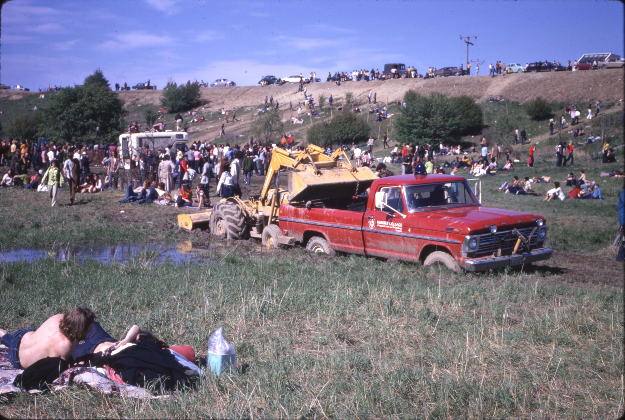 A pickup truck sits in a field with farm machinery behind it. A large number of people are gathered nearby.