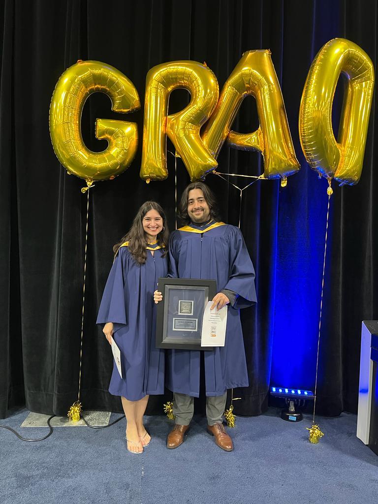 Two people wearing convocation gowns stand beneath balloons that spell out grad.