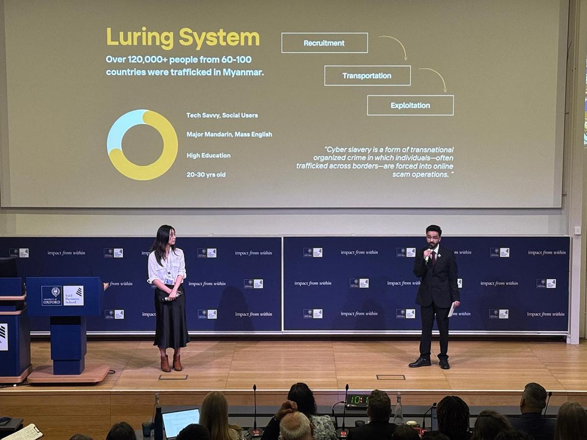 Two people stand on a stage holding microphones with a screen with a presentation behind them. 
