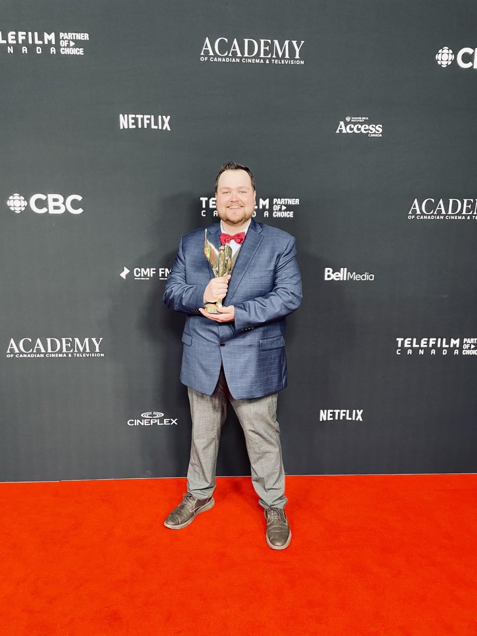 A person holding an award stands on a red carpet in front on a backdrop with writing on it.