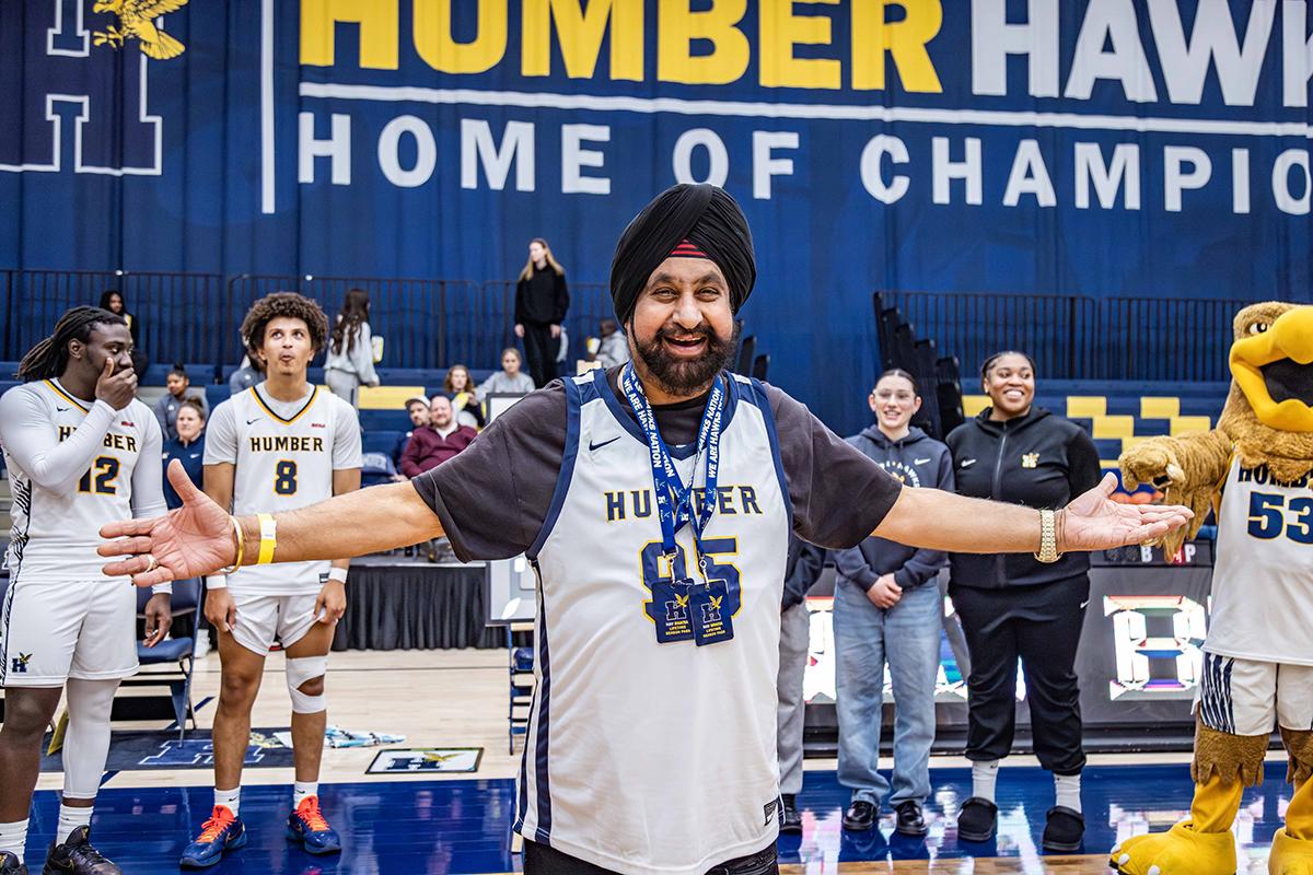 A person wearing a Humber Hawks jersey stands with their arms outstretched as other people in the background clap.