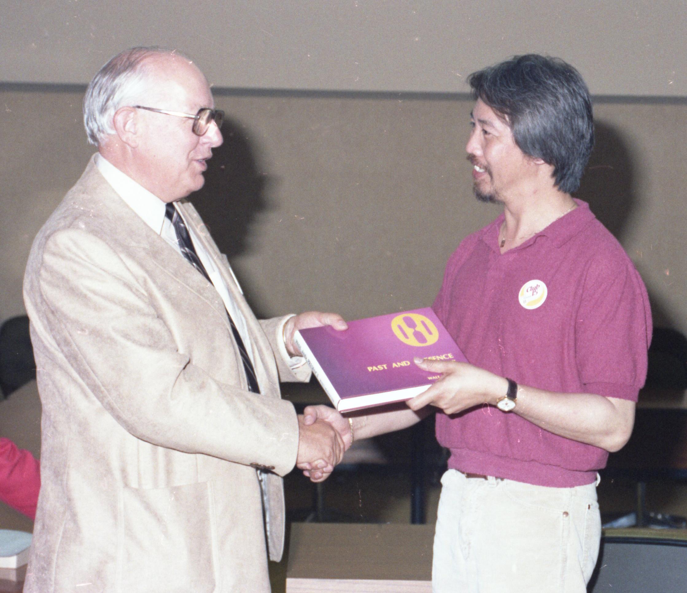 Two people shake hands while one is presented with a book.
