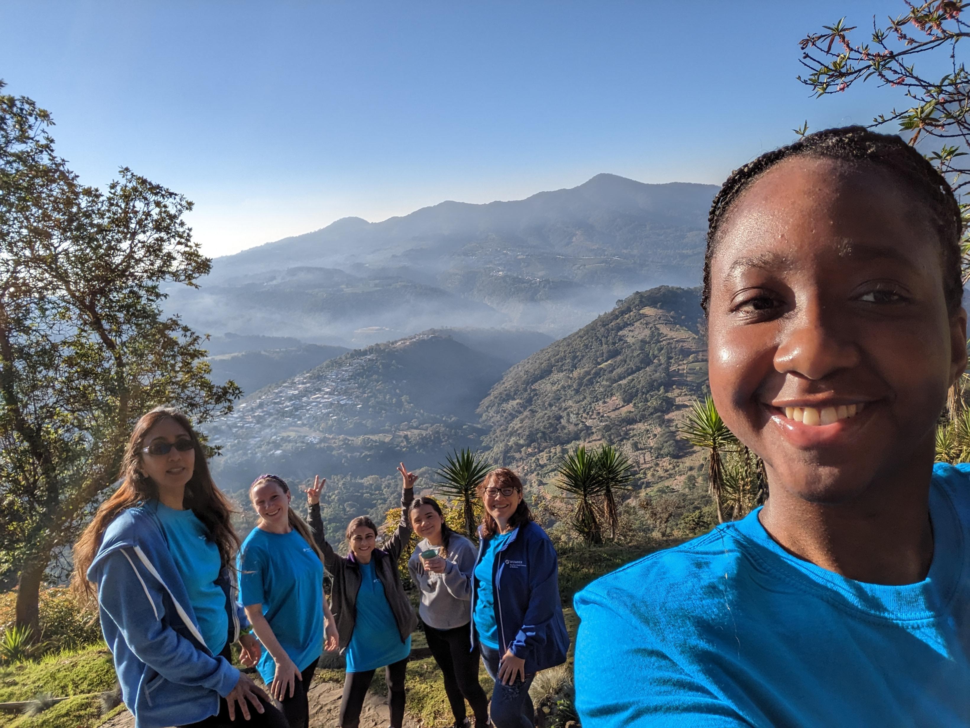 Six people take a selfie with a picturesque mountainous area behind them in the background.