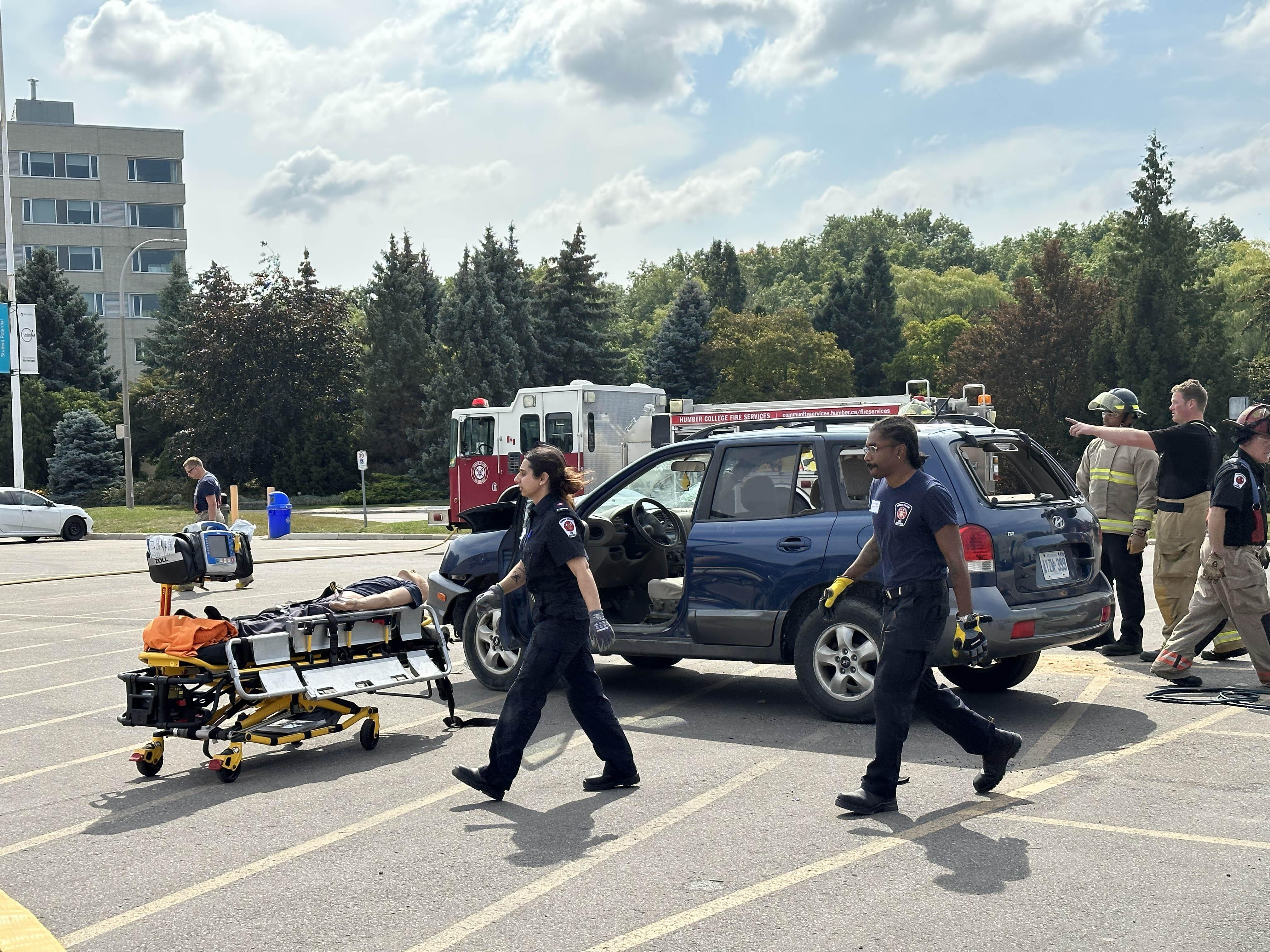 Students wearing paramedic outfits walk towards a stretcher with a training duty strapped to it.