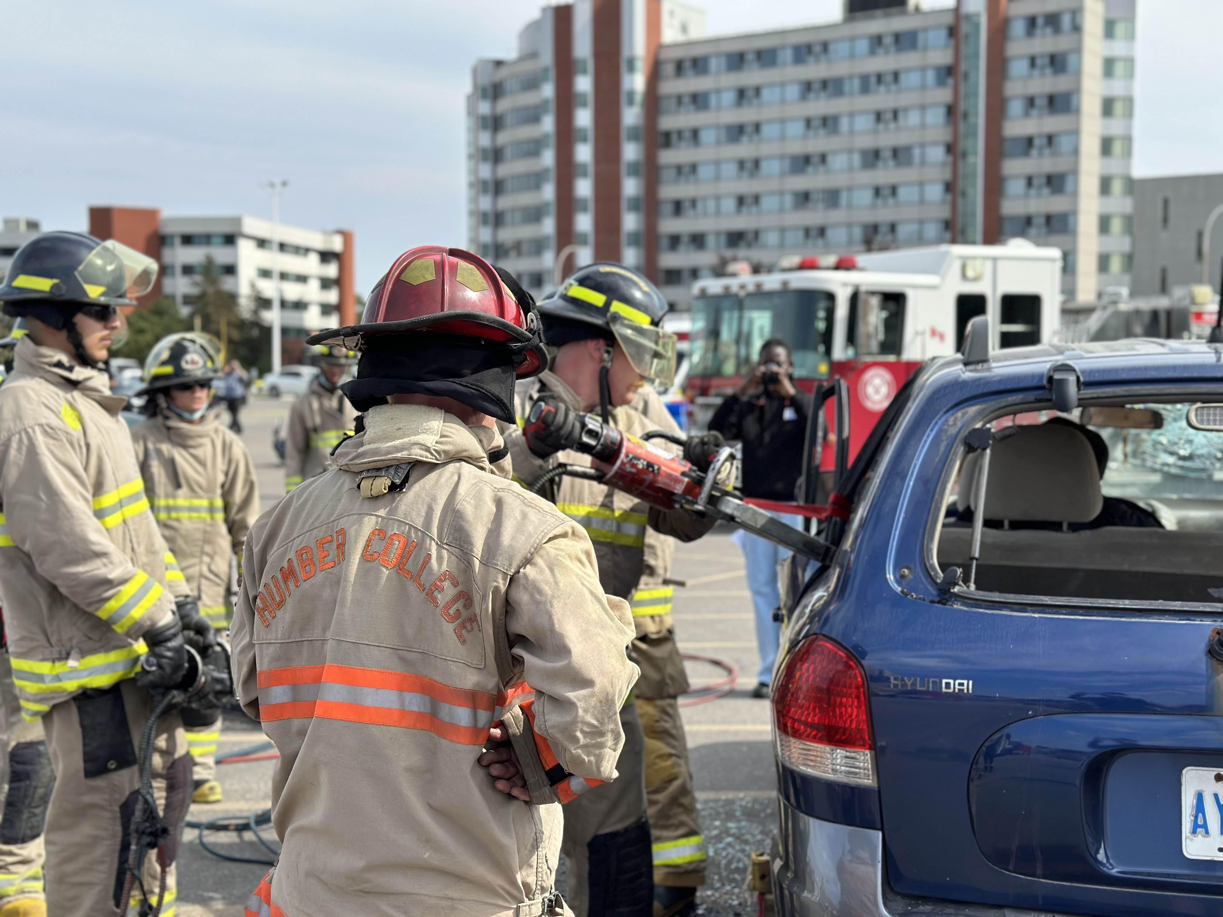 Students wearing firefighter equipment watch as anther person use the 'Jaws of Life' to open the door of a vehicle.