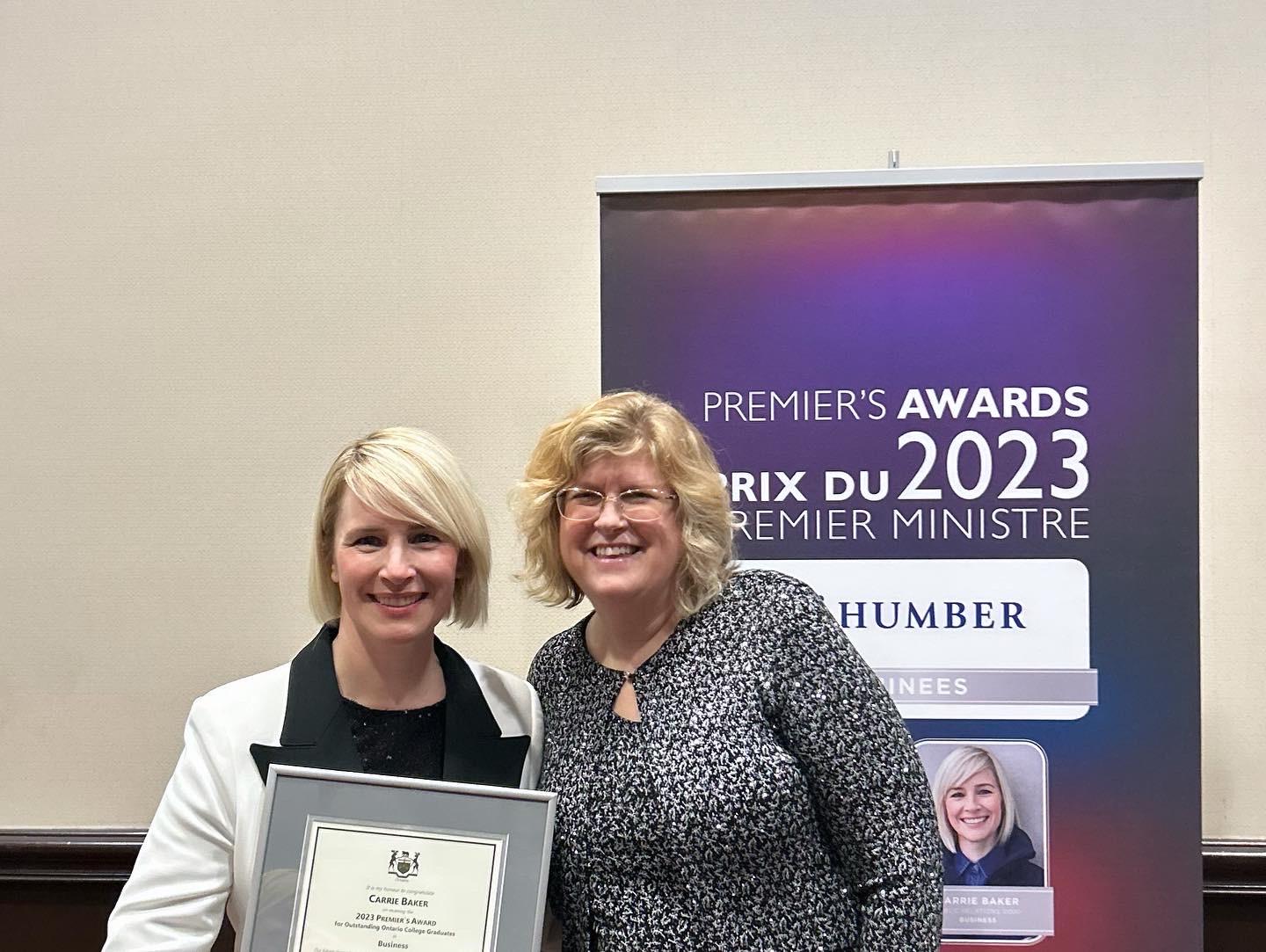 Two smiling people pose for a photo in front of a banner that reads Premier’s Awards 2023. One is holding a framed document.