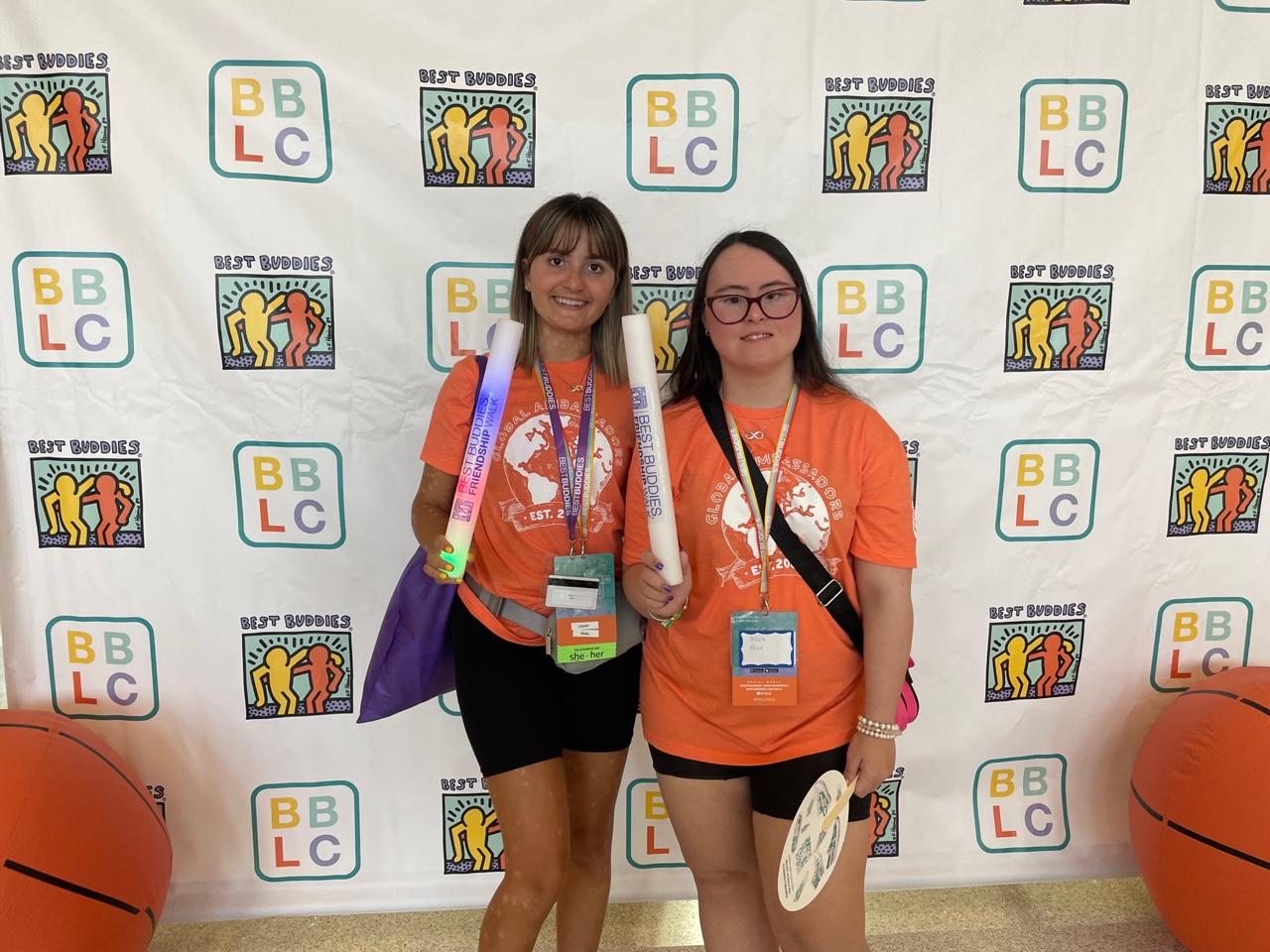 Two people holding inflatable tubes stand in front of a white backdrop that reads Best Buddies.