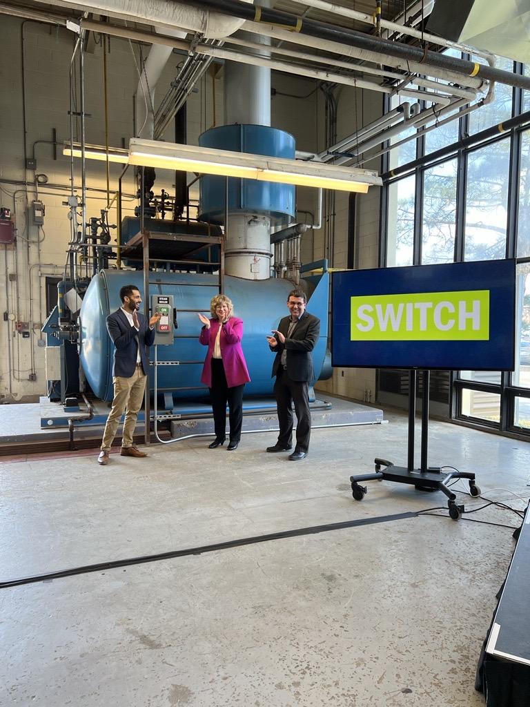 Three people standing in front of a large mechanical device clap. A screen beside them reads SWITCH.