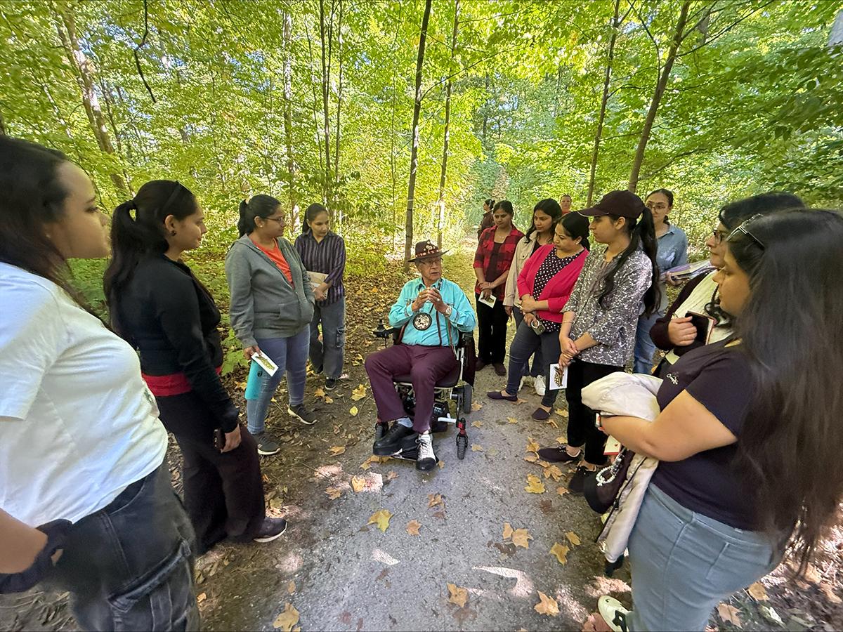 The Humber Polytechnic community is encouraged to participate in a self-guided StoryWalk at the Humber Arboretum. A group of people stand in a forest listening to another person speak.