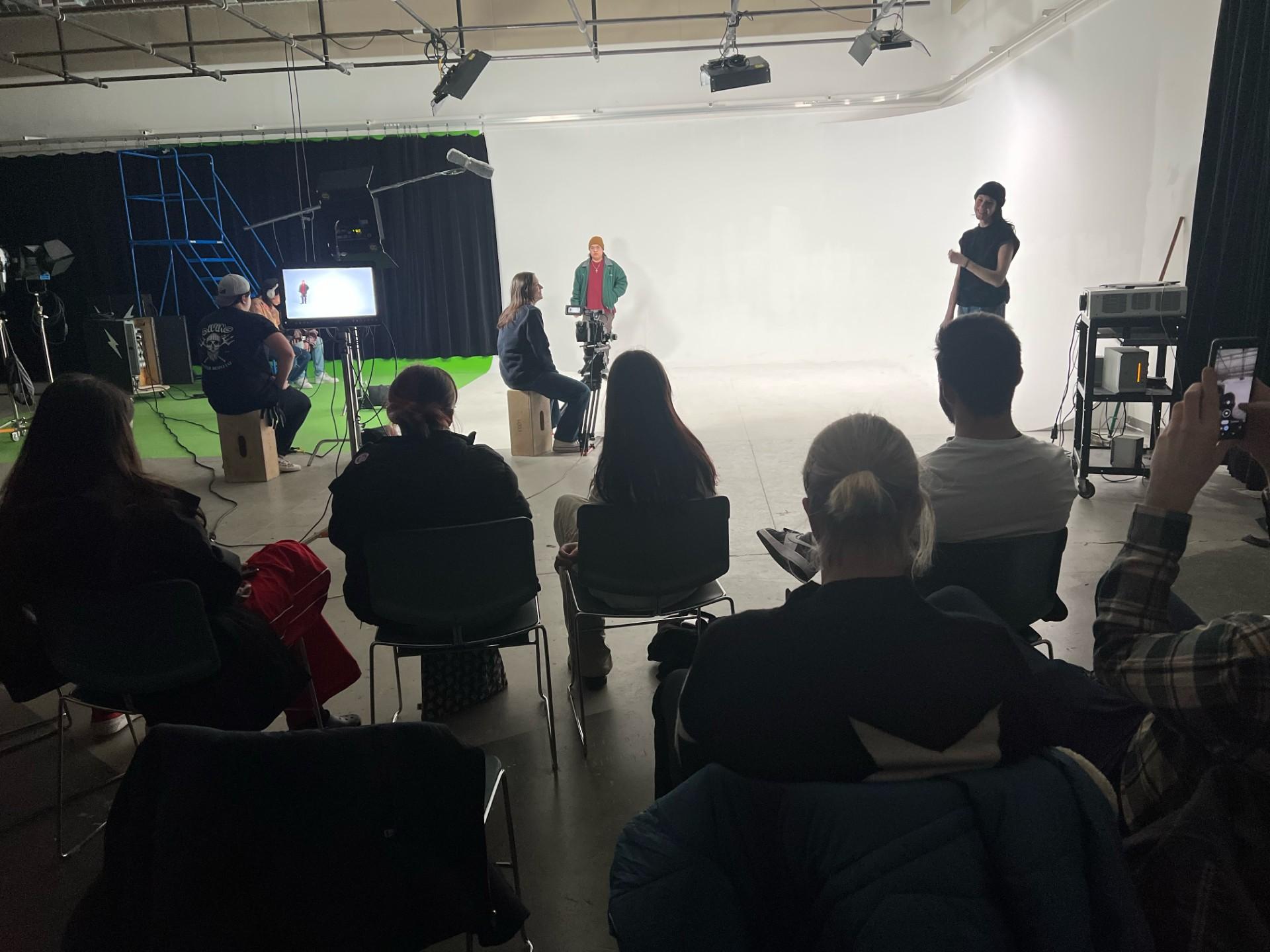 People sit in a studio and watch as others are filmed standing in front of a white screen.