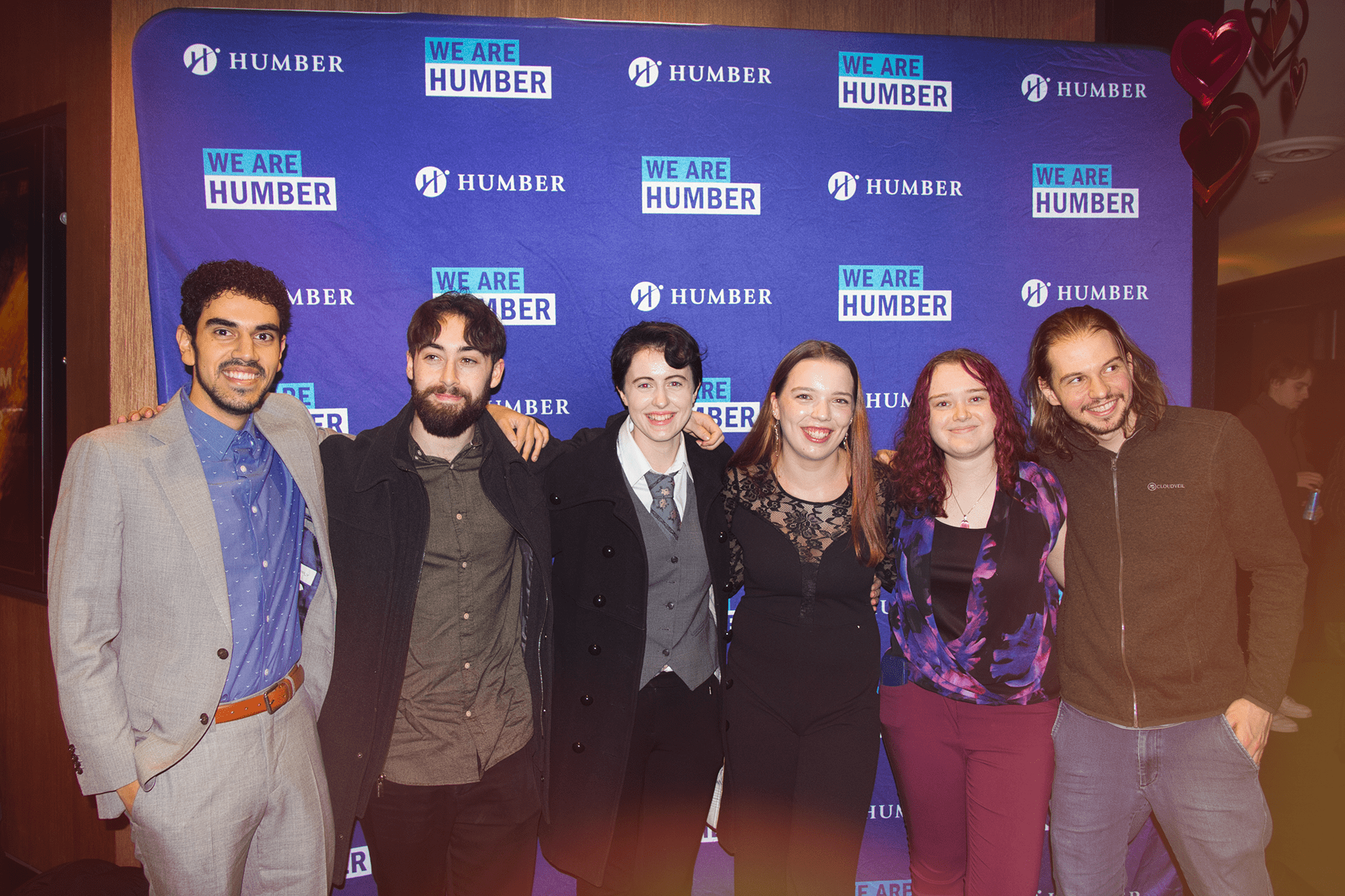 A group of six smiling people stand together in front of a blue backdrop with the Humber College logo on it.
