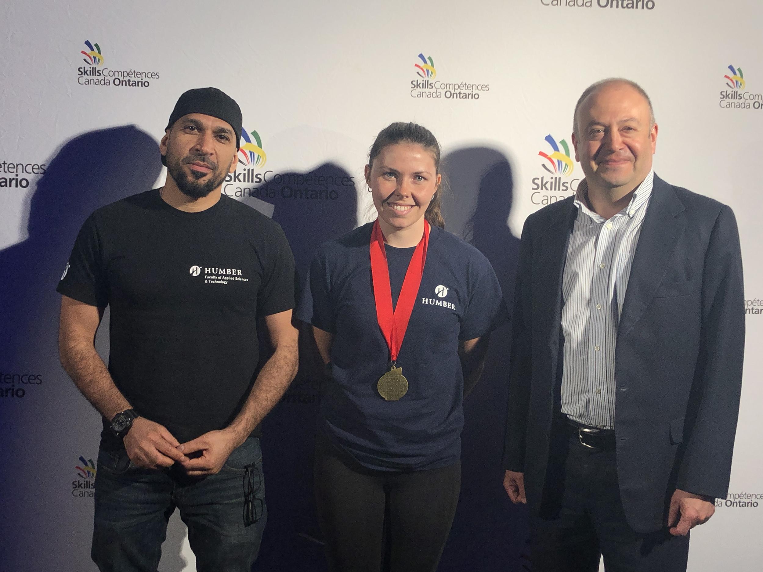 A smiling Humber College student wearing a gold medal around their neck is flanked by two other people.