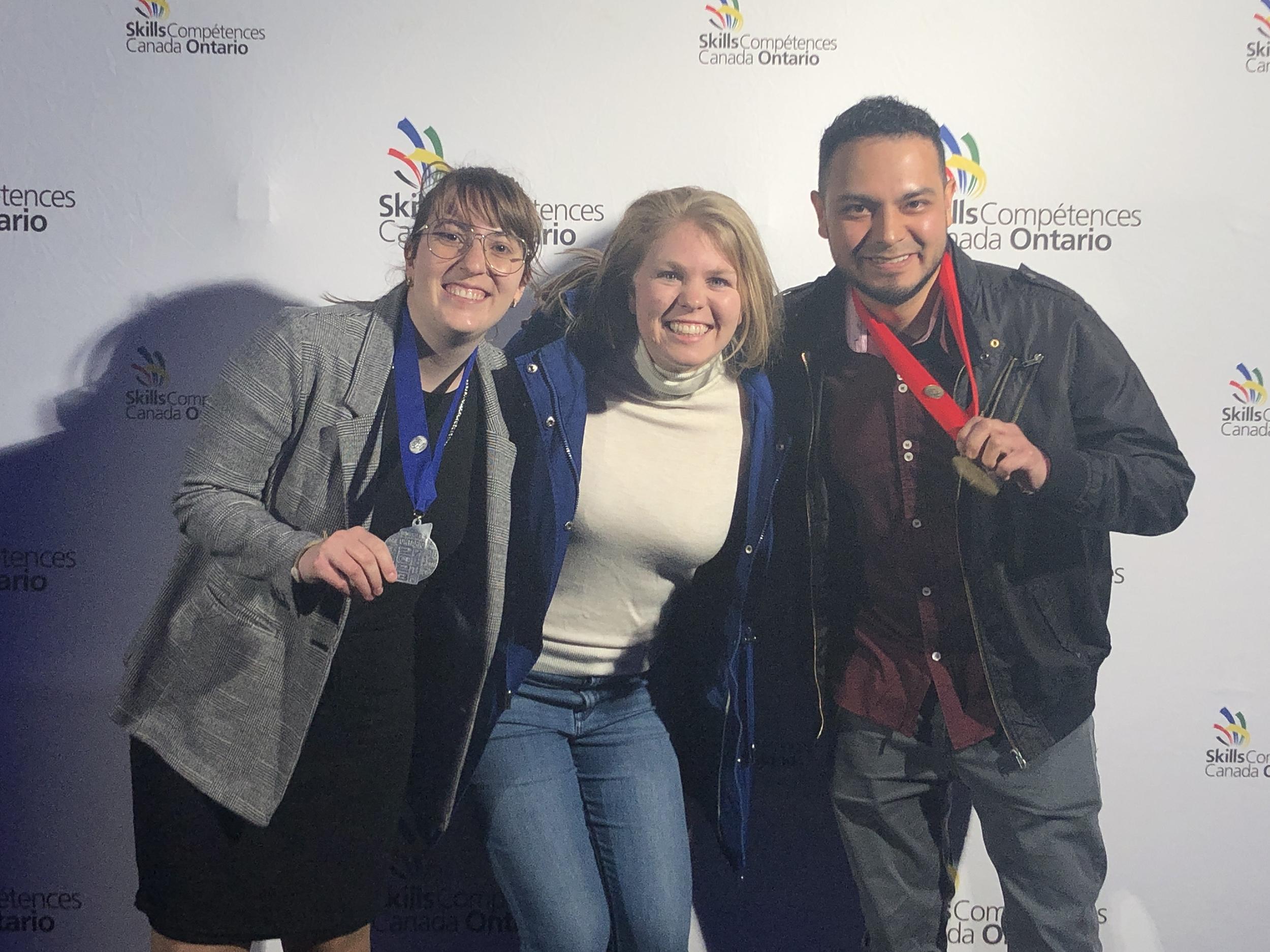 Three smiling people pose for a photo. One person has a gold medal around their neck and the other has a silver medal.