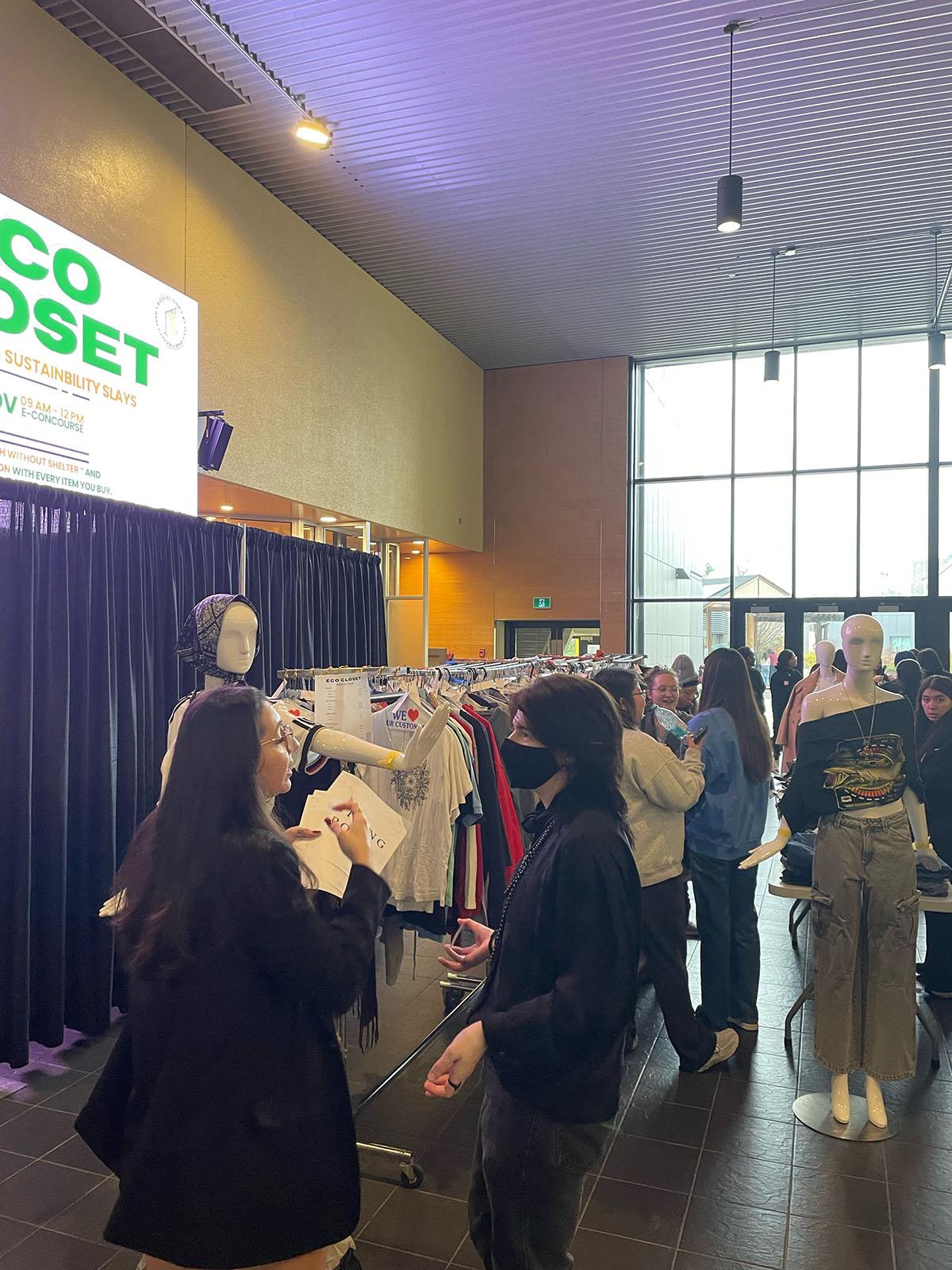 Two people have a discussion while other people peruse racks of clothing.