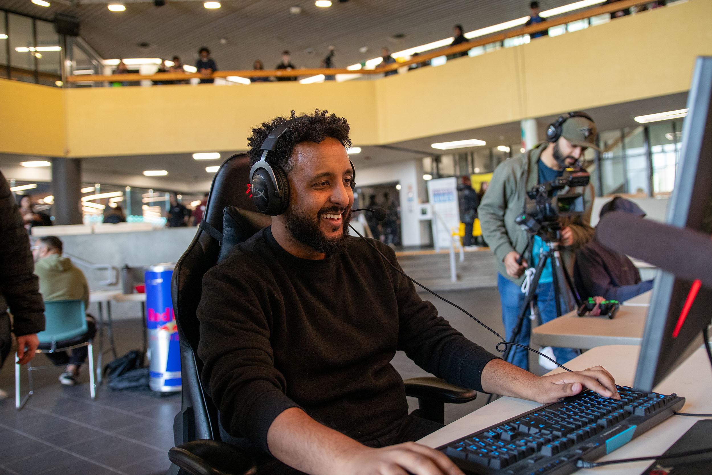 A smiling person wearing a headset sits at a computer with their hands on the keyboard.
