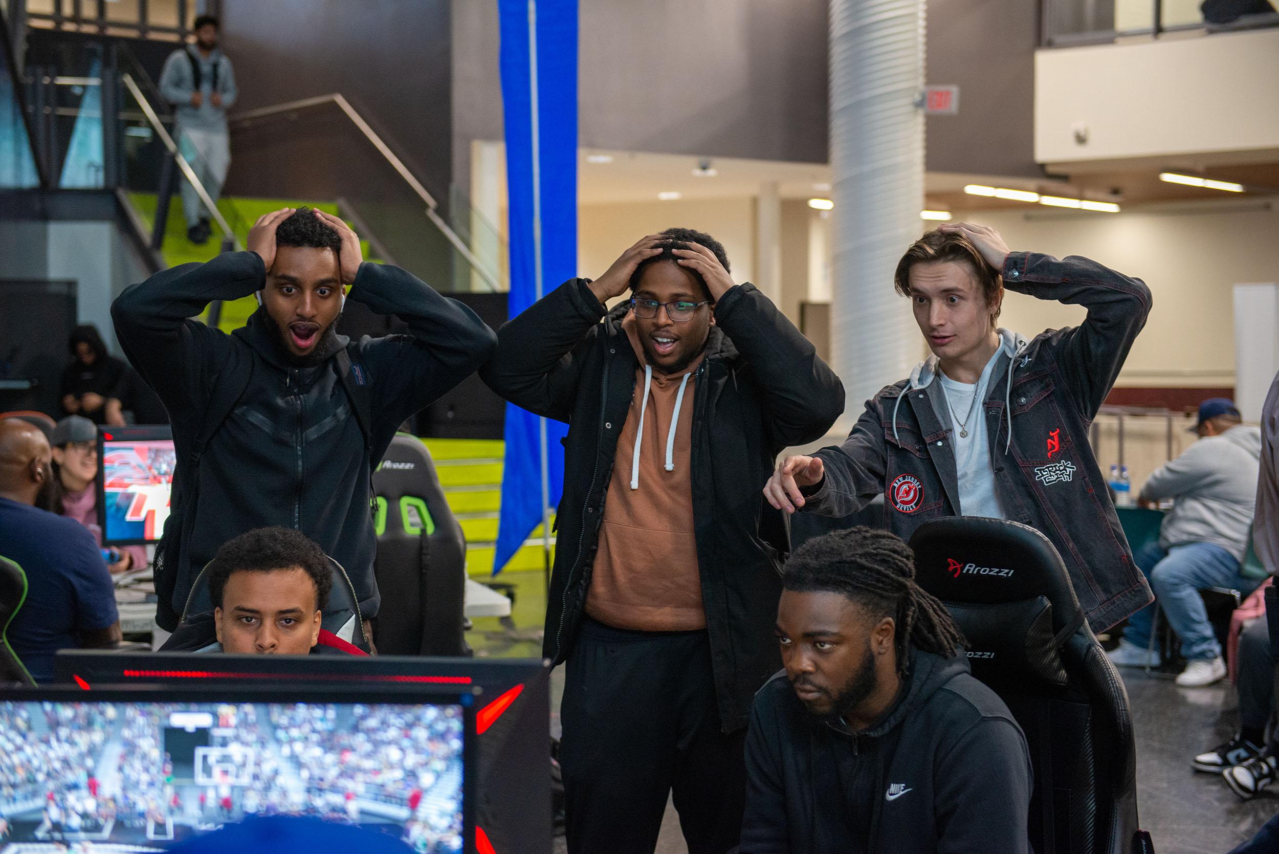 Three people put their hands on their head in astonishment while watching others play video games.