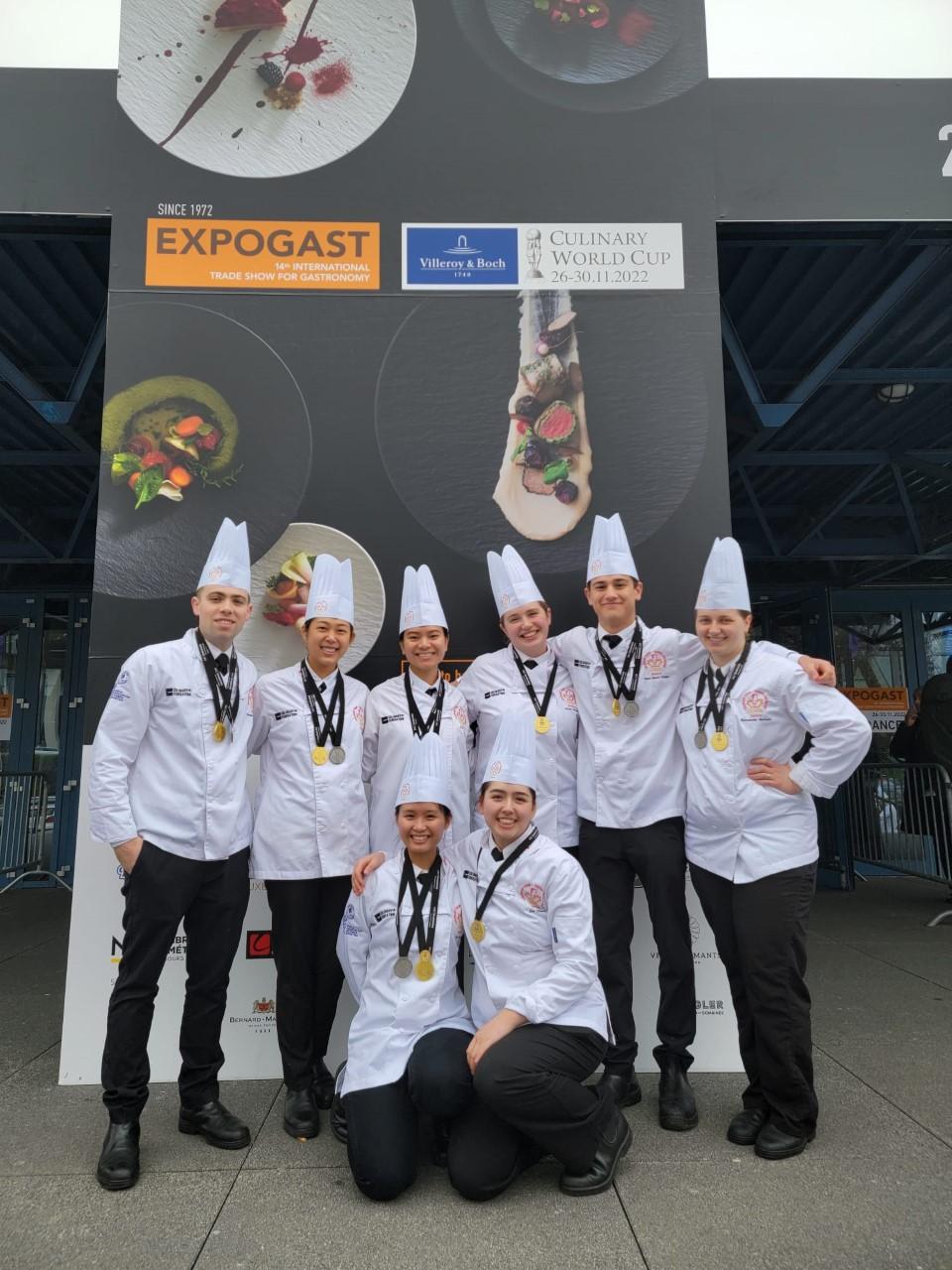 A group of eight people wearing chef’s uniforms with gold and silver medals around their necks.