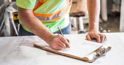 A person wearing a high-visibility yellow shirt hunches over a clipboard with a pencil in their hand