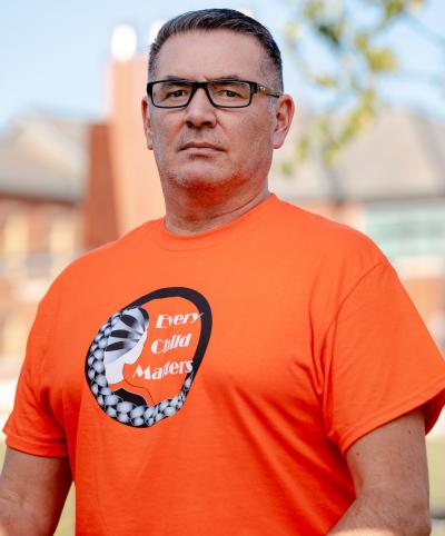 Jason Seright is wearing an orange shirt and sitting outside on a bench with trees in the background.