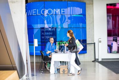 A person walks past another person sitting at a table in front of a digital sign that reads Welcome, Humber Alumni Weekend 2022.
