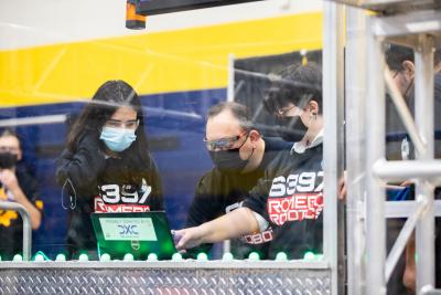 Students stand behind plexiglass as they control their robots in the competition area