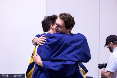 Two people wearing graduation gowns embrace with a hug.