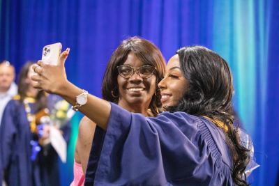 Two people, one wearing a graduation gown, smile and look into the camera as they take a selfie.