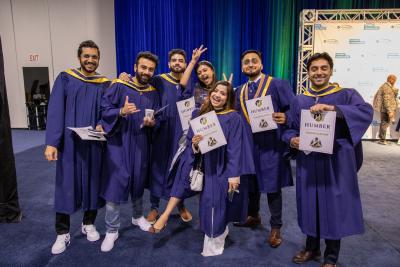 A group of students wearing graduation gowns smile and pose for a photo while holding up signs that say Humber and congratulatio