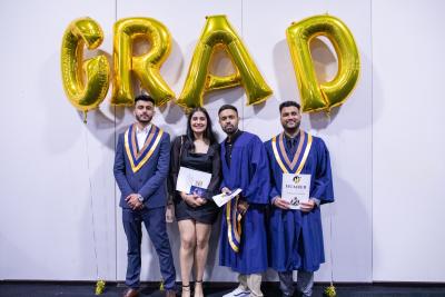 Four graduates smile and pose for a photo while standing underneath a balloon sign that reads ‘grad’.