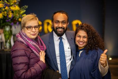 Three people, one of whom is a wearing graduation robes, embrace and smile for a photo.