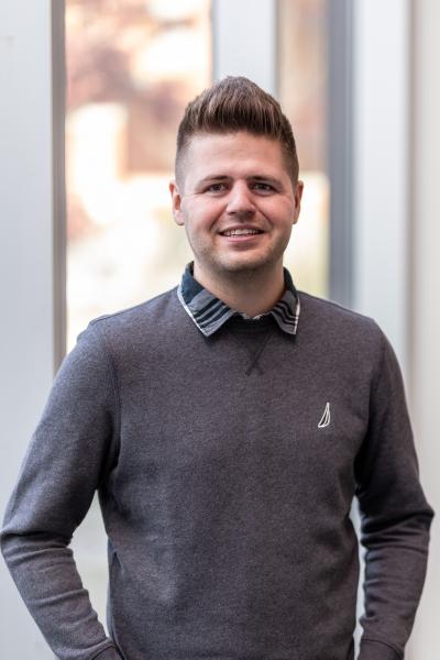 Casey Norris stands, hands on hips, in front of a window wearing a grey sweater and a collar, smiling