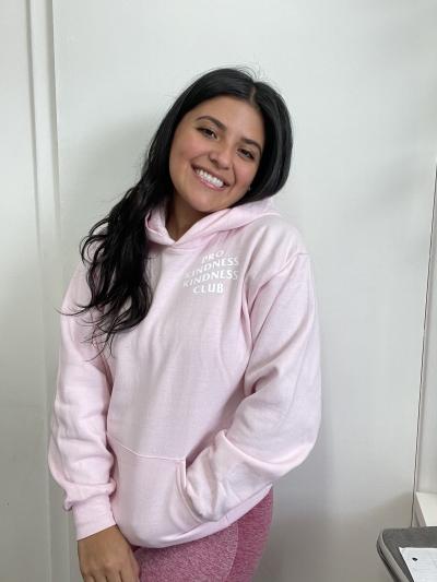 Camila Ruiz Tacha has long brown hair and one hand in the pocket of her pink sweatshirt and smiles, head tilted to the side.