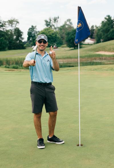 A person stands next to the pin on a golf course while holding a ball and a golf club.