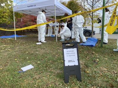 Students wearing white jumpsuits document a simulated gravesite. A sign reads Academic Simulation in Progress.
