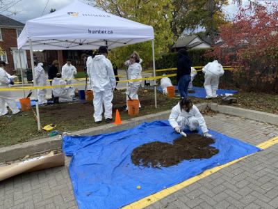 A student sifts through dirt on a blue tarp looking for evidence while other students use shovels to dig up earth.. 