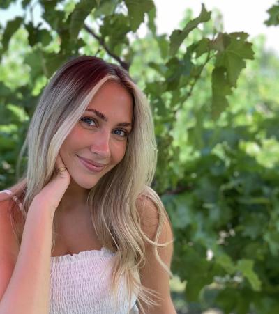 Maddison Gerrie smiles slightly in a headshot with her hand resting near her face. There is green foliage behind her.