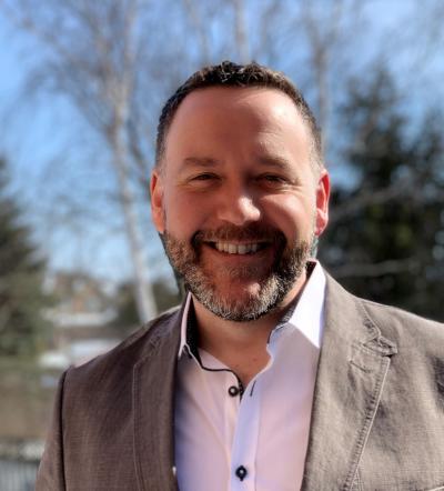 Chad Crago is smiling in front of a background of trees and blue sky. He has a short beard and wears a button down and jacket