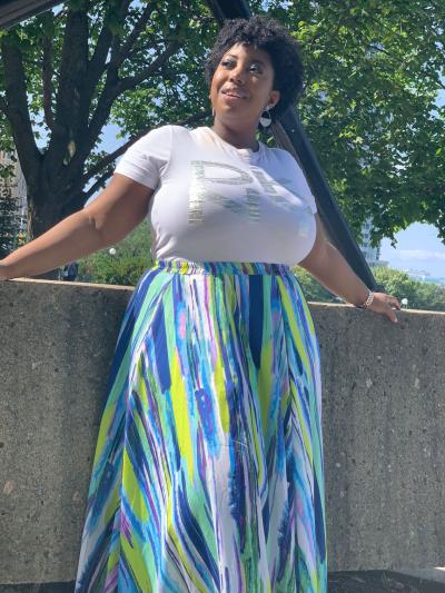 Akaila Reid leans on a short cement wall, looking sideways up at the sun and smiling. She wears a white top and colourful skirt