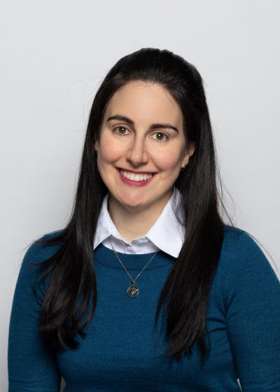 Program coordinator Erin Mandel-Shorser smiles in a headshot, wearing a collared shirt and green sweater in front of a white wal