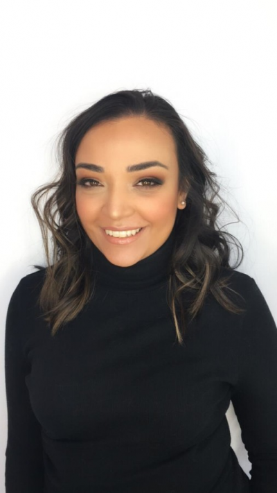 Jessica Harwood smiles in front of a white background. She has long brown hair in curls and is wearing a black turtleneck