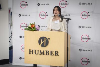 Humber student Nathalie Gomez speaks into a microphone while standing at a podium that has the word Humber on it.
