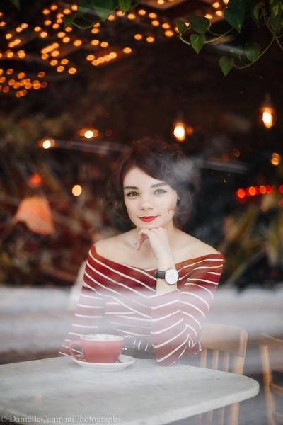 Laura Anglade is seen through a window at a coffee shop, posed in front of a cup and saucer with her hand under her chin